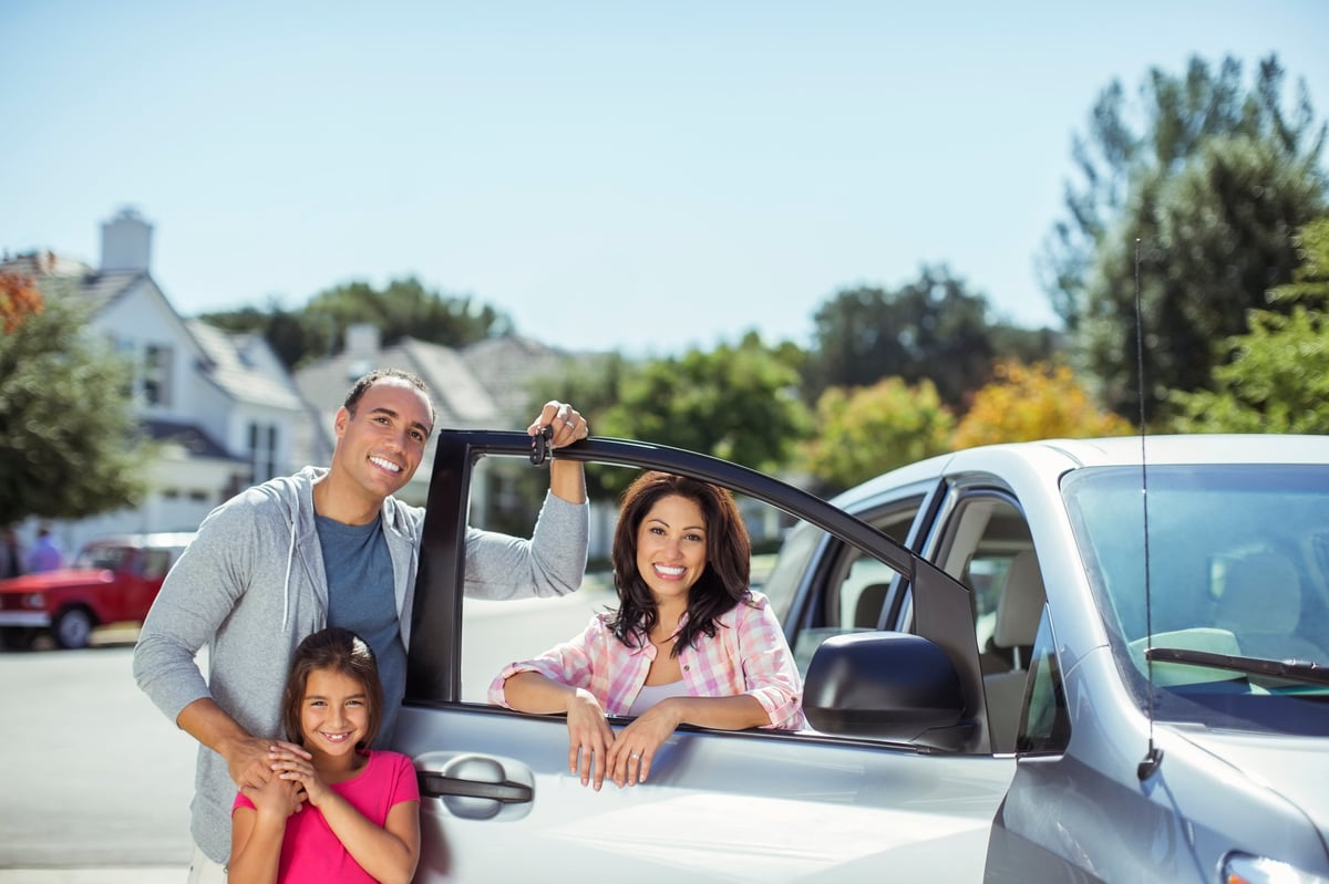 Family of three smiling near a parked car in a suburban neighborhood on a sunny day.